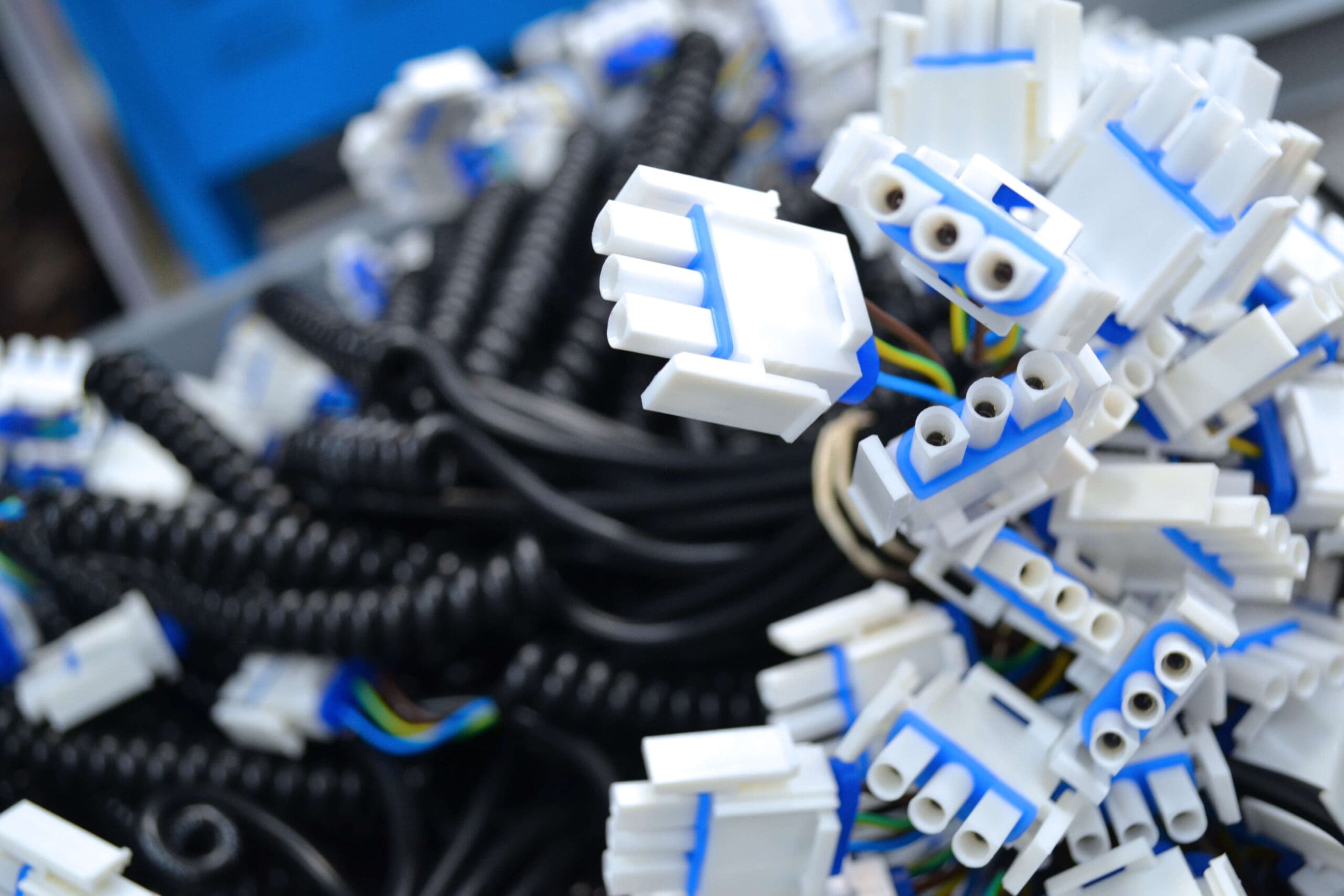 The image shows a large number of electrical connectors and coiled cables. The connectors are primarily white with blue accents, featuring multiple sockets, likely designed to accommodate spade or similar connectors. The black coiled cables suggest flexibility and potential use in environments where length variability is an advantage, like in telephone handsets or other electronic devices that require movement. The background is blurred with hints of blue, focusing the attention on the connectors in the foreground. The abundance and disarray of the connectors indicate a storage or supply setting, perhaps in an electronics workshop or manufacturing facility.