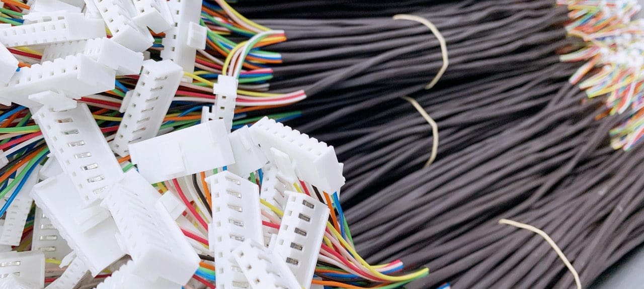 The image shows a bundle of black cables on the right, neatly organized and tied together, fanning out into individual multi-colored wires. These wires are connected to white plastic connectors, which are in a disordered pile on the left. The connectors appear to be ribbon cable connectors, commonly used in electronics to connect multiple data lines or power lines between two points with a single plug-and-socket connection. The array of colors within the cables typically follows a standard electronic color code to identify each wire's purpose. This setup suggests a mass production or testing environment for electronic components.