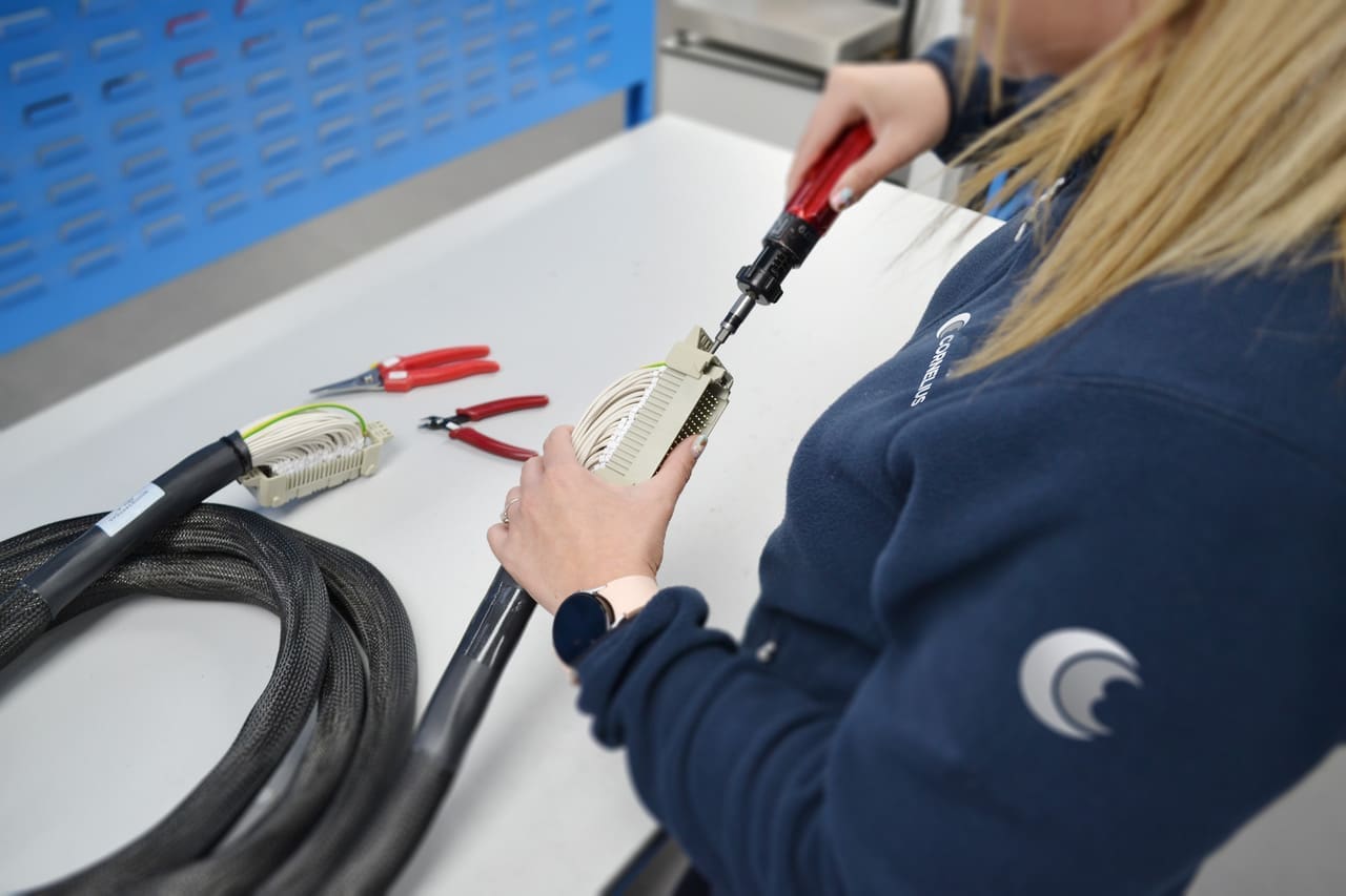 An employee at Cornelius Electronics is assembling a complex cable connector with precision tools on a workbench. Pliers and another cable lie beside her, and she's wearing a branded sweatshirt with the company logo.