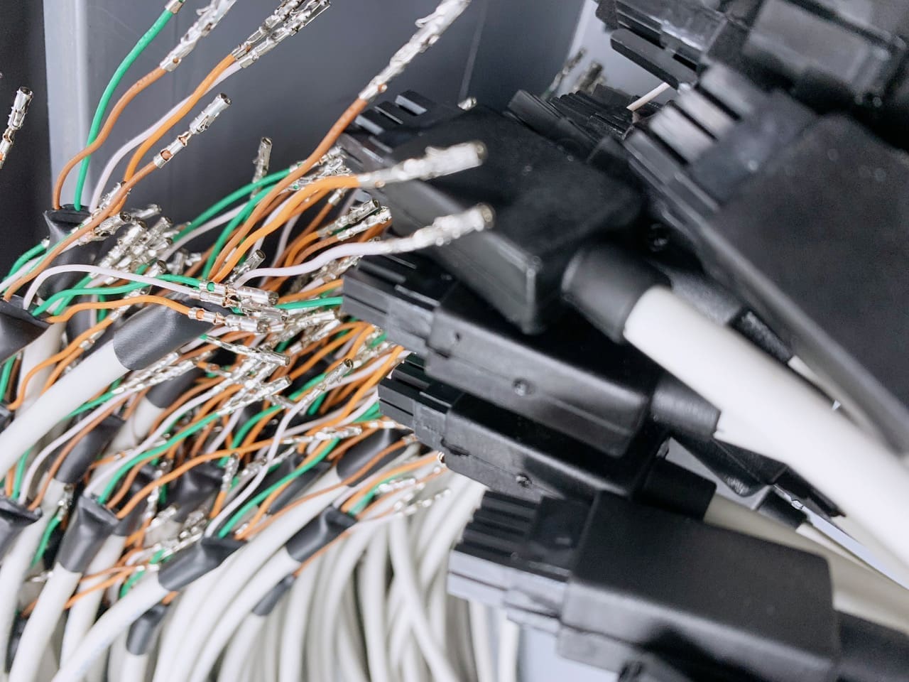 The image shows a close-up of a cluster of cables and connectors. We see a mix of shielded and unshielded wires, with various colours like orange, green, and white. Some of the wires are stripped at the end, exposing the metal conductors. These are typically used for making electrical connections in a system. A number of black connectors are visible, suggesting that these cables are part of a larger assembly or network of connections, possibly within a data center, telecommunications system, or complex electronic equipment. The focus is slightly blurred, adding a sense of depth and complexity to the array of connections.